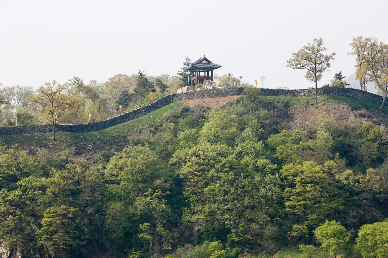 A low stone fortress wall runs along a wooded ridge with a red-pillared pavilion on top