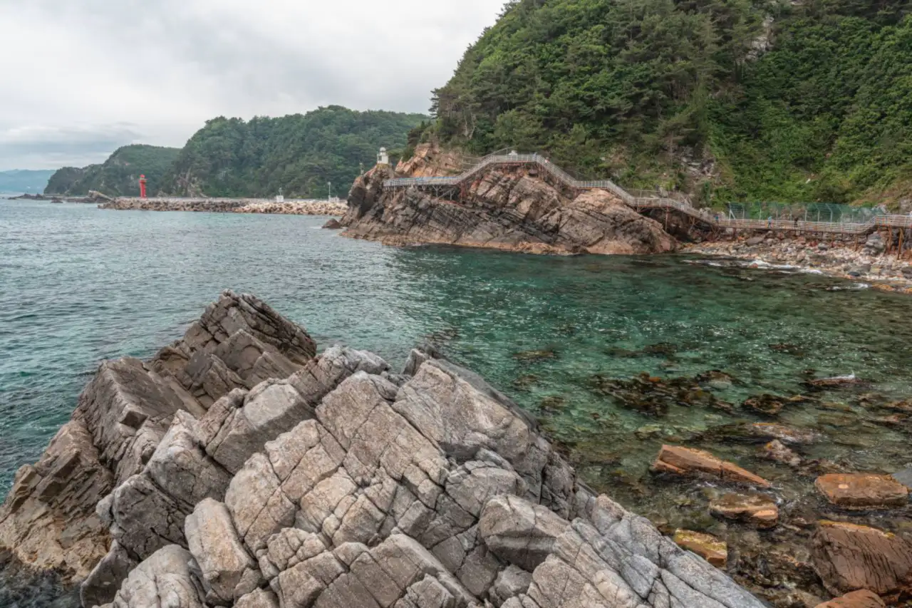 Layered grey rocks in the foreground, with the steel boardwalk along the cliff and a red breakwater lighthouse in the distance