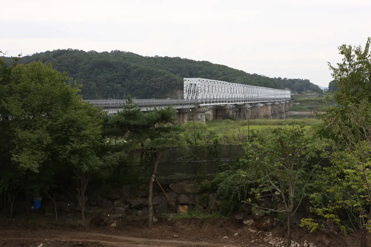 A white truss railway bridge over the Imjin River with wooded hills behind
