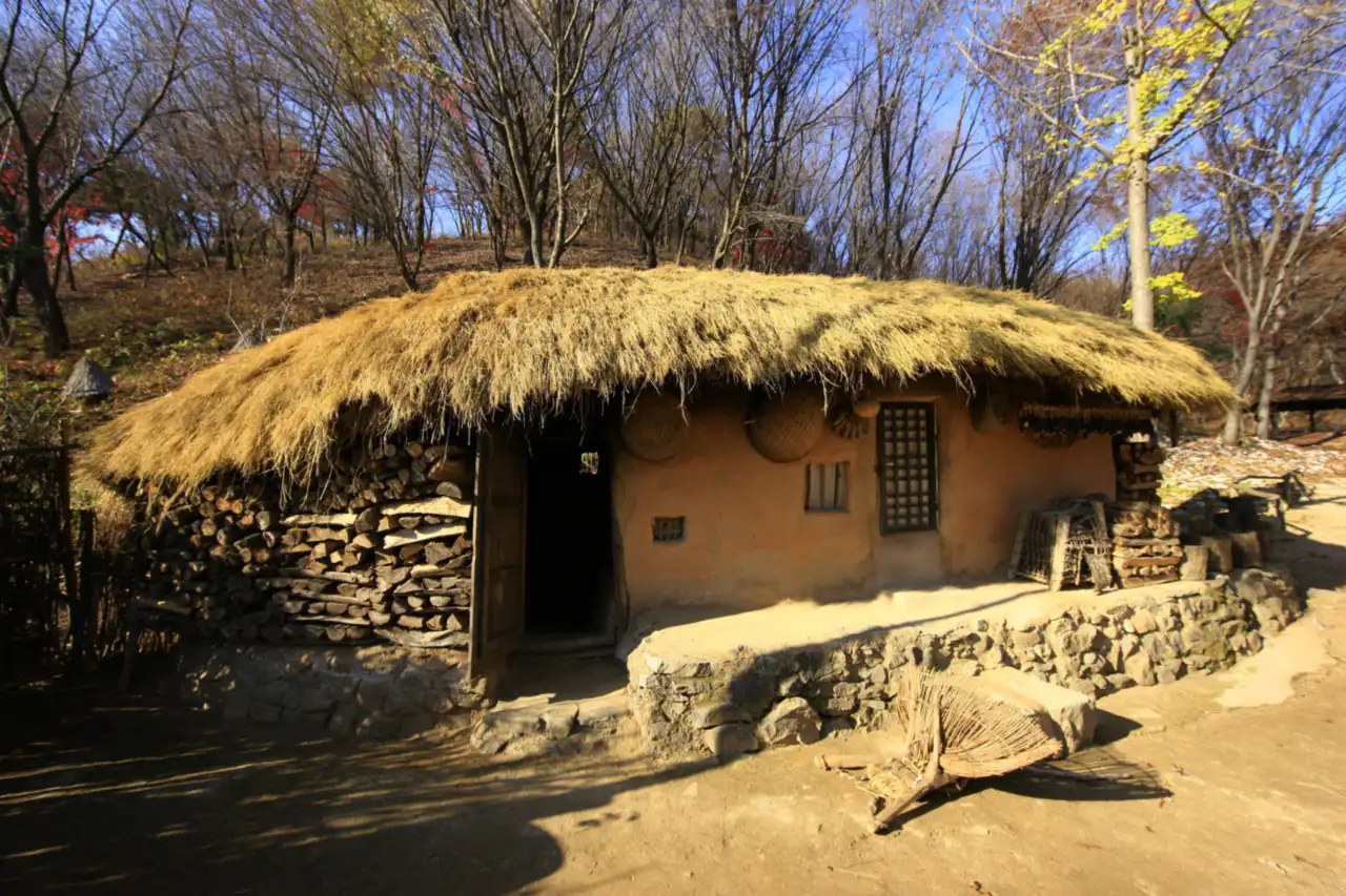 A thatched-roof clay house with stacked firewood against autumn woods