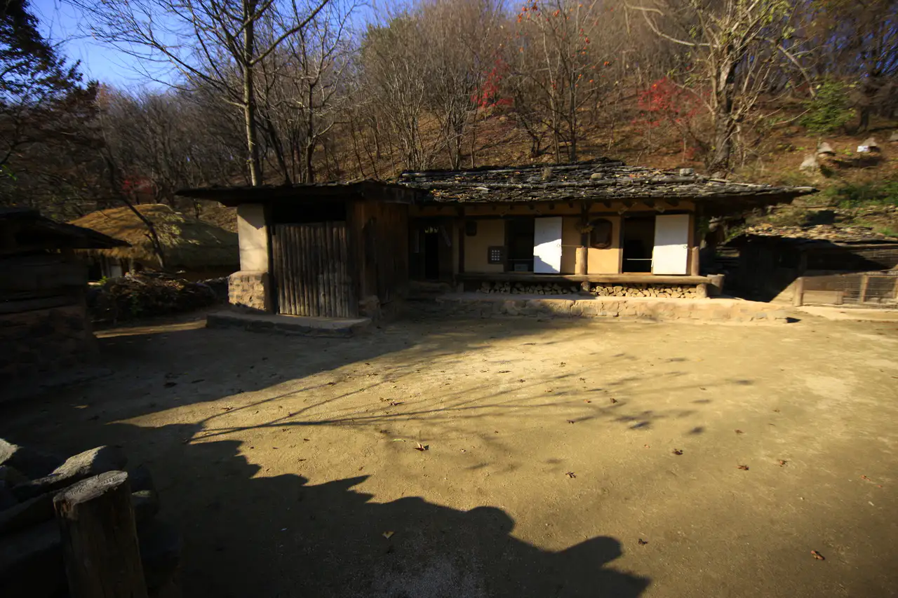 A clay-walled house and dirt yard below a wooded hillside