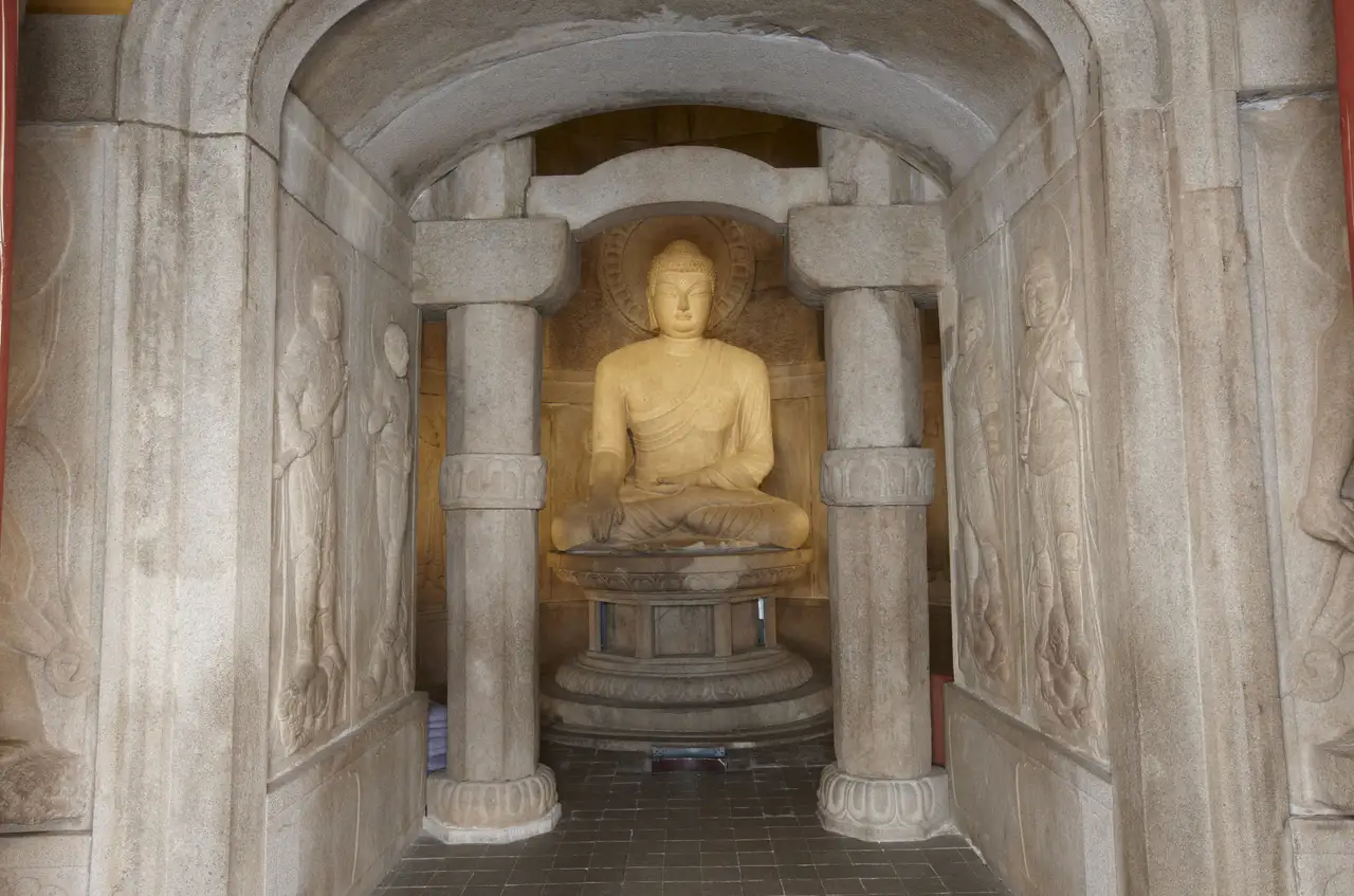 At the end of a stone passage carved with guardian kings, the lit main Buddha sits between two octagonal stone pillars