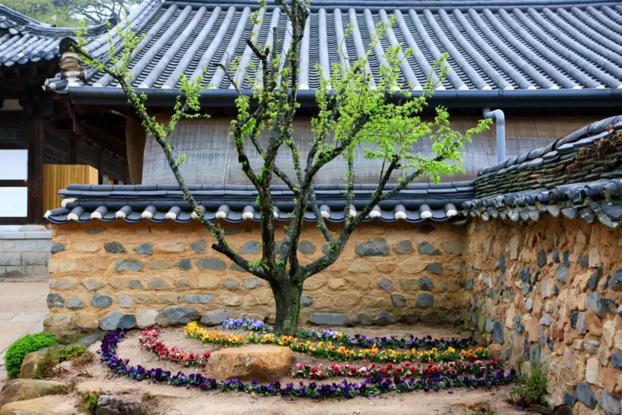 Spring flowers planted in a spiral beneath a tree before a stone wall and tiled roofs