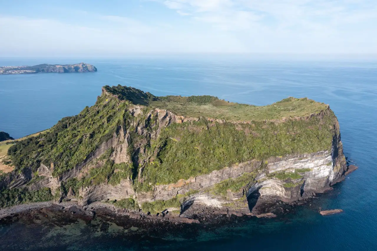 Aerial view of Seongsan Ilchulbong: steep cliffs and the grassy crater rim rising from a blue sea