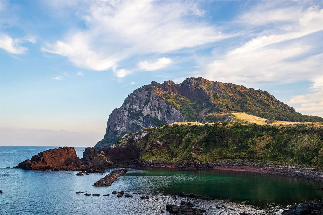 The rocky peak of Seongsan Ilchulbong above red volcanic rocks and a calm cove