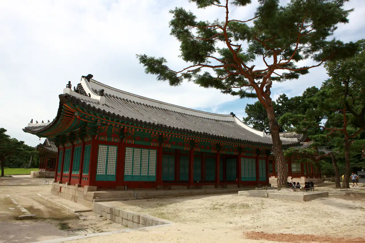 A red-pillared hall with green lattice doors on a low stone base, a pine spreading beside it across a sandy courtyard