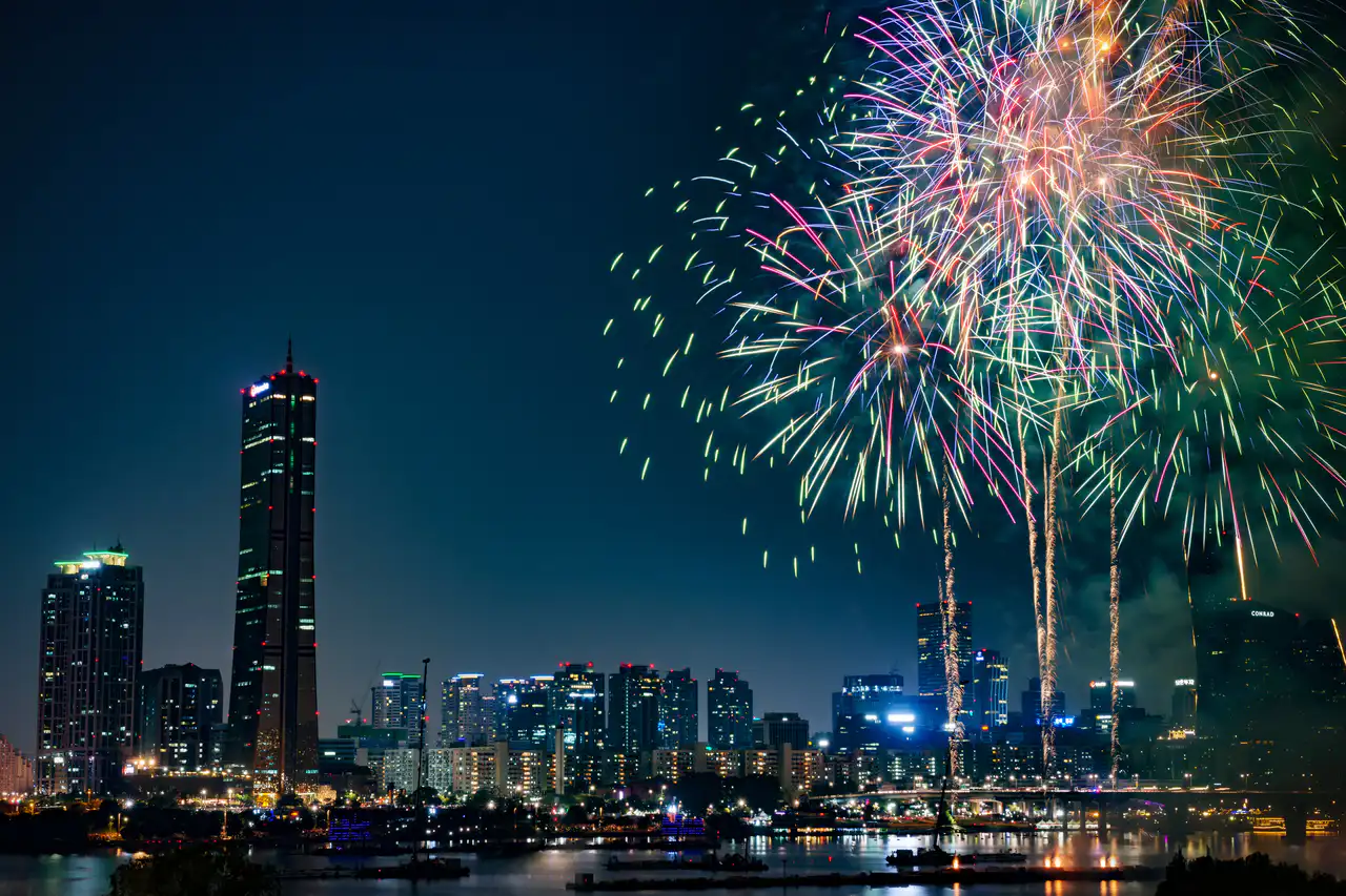 Fireworks bursting over Yeouido at night, the 63 Building and city lights reflected in the river